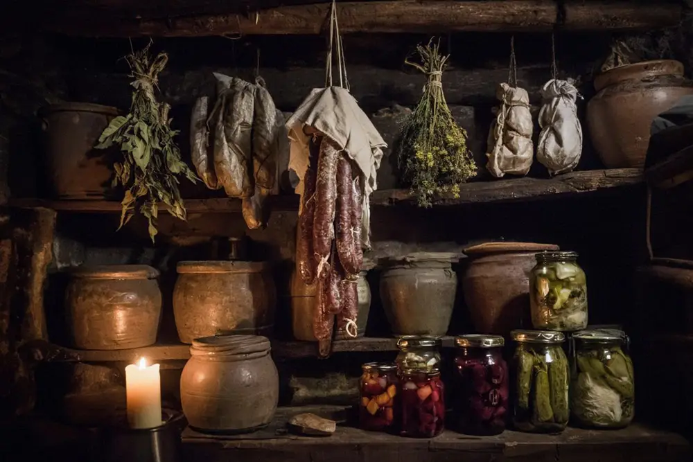 A dark cellar shelf lined with ceramic crocks, hanging dried herbs, salt-cured meats wrapped in cloth, and glass jars of preserved vegetables