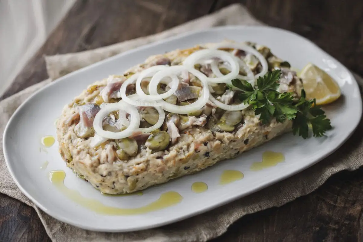 A oval loaf of mashed broad beans and herring on a long ceramic plate, drizzled with olive oil, garnished with sliced onion and parsley